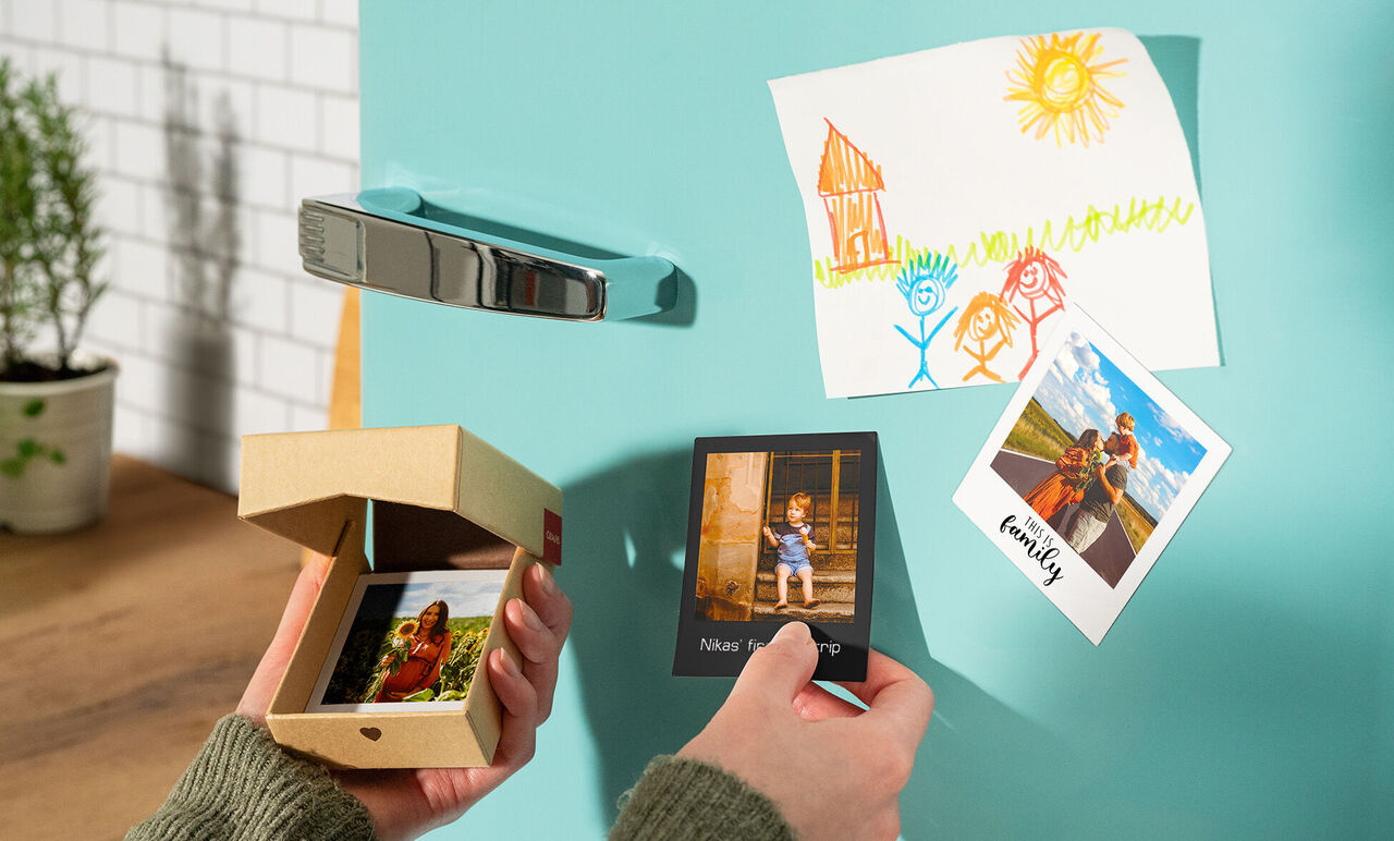 A person opens a small box with photos and hangs a picture on the fridge. A family photo and a colorful drawing are also seen on the fridge.