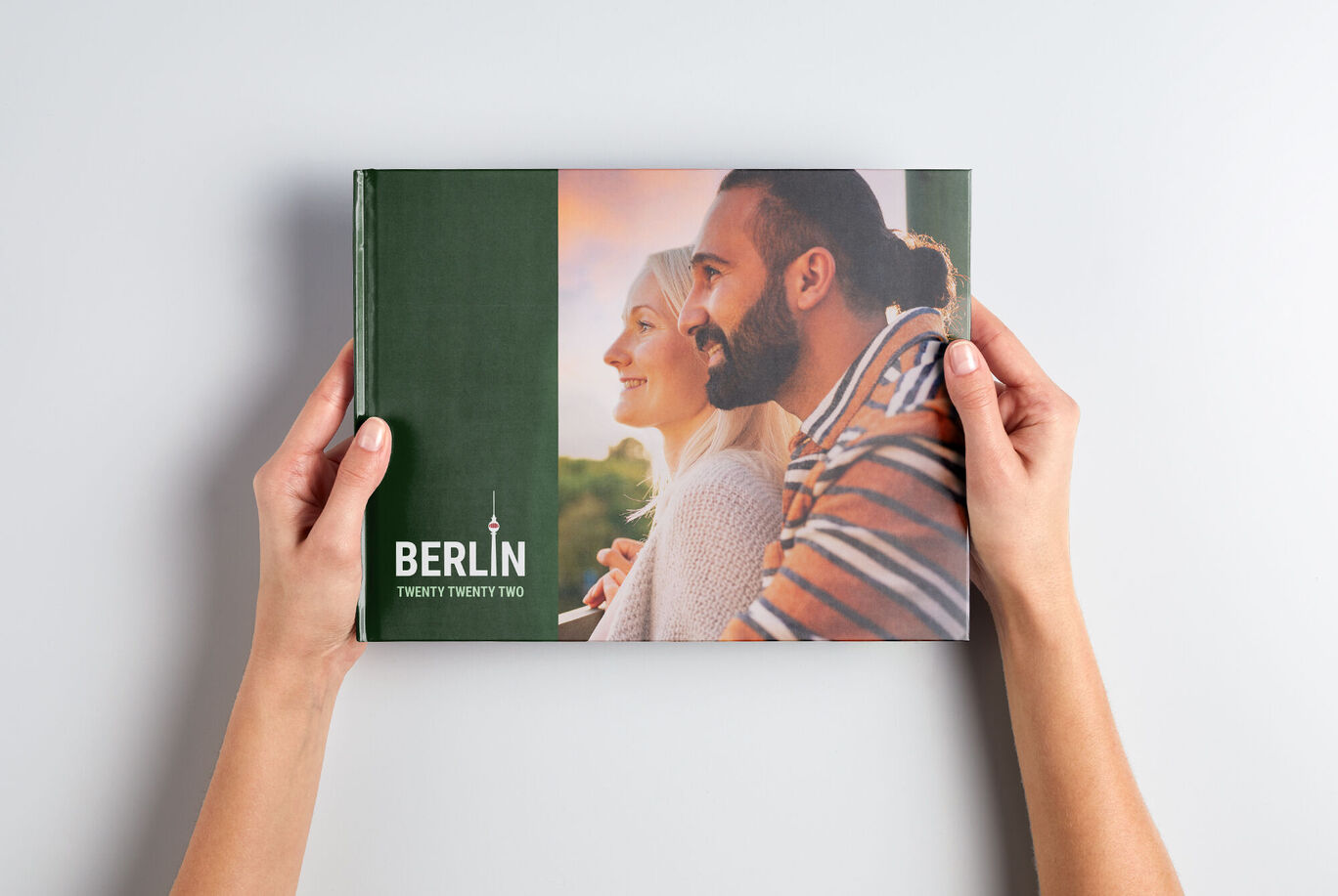 Hands hold a CEWE photo book titled 'Berlin Twenty Twenty Two' against a light background