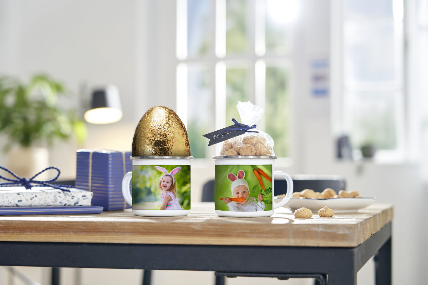 Two personal mugs with memory photos on a table, next to a golden egg and sweets.
