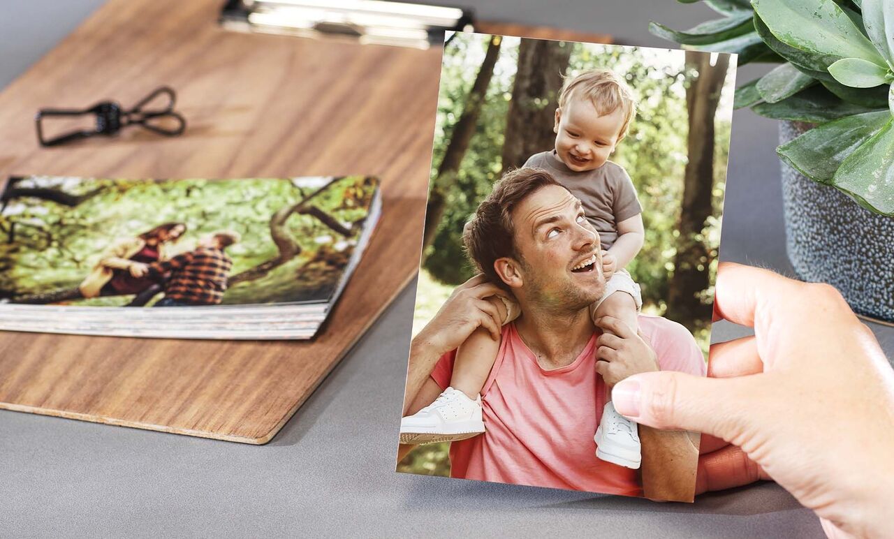 A hand holds a photo of a father with his child on his shoulders. More photos are on a table in the background.