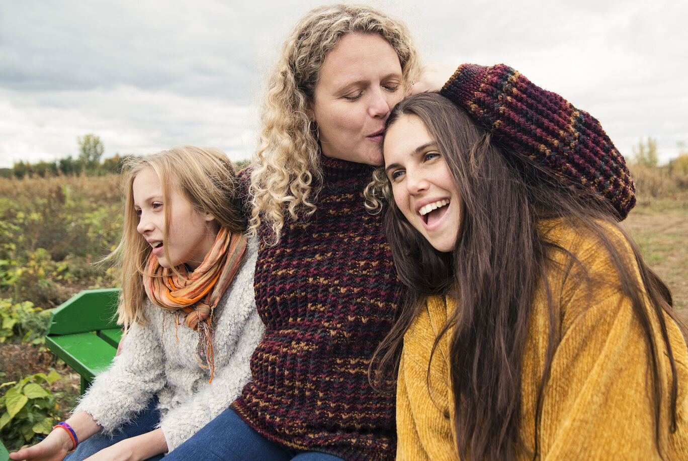 Three women sit together outdoors in warm sweaters and smile, in a field surrounded by nature.