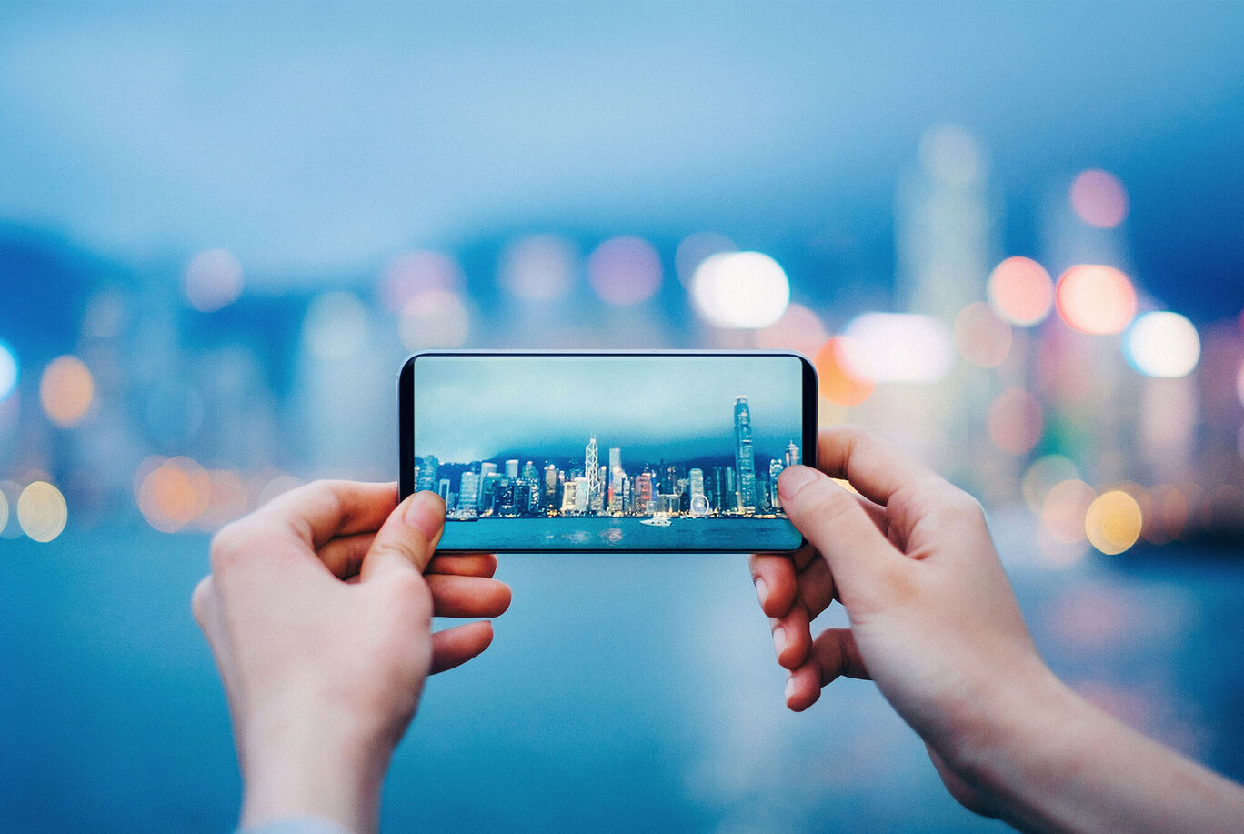 Person holding a smartphone photographing the harbor with illuminated buildings in the distance