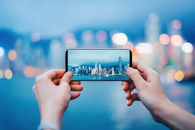 A person holds a smartphone displaying a city view. The skyline shines at dusk.
