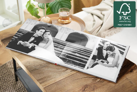 A beautifully designed CEWE photo book lies on a wooden table, showing black and white photographs of a wedding couple on the steps of an elegant venue.