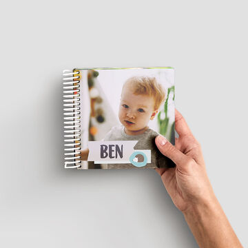 A lovingly designed CEWE photo book is held by a hand. The title on the book is "Ben" and it shows a cheerful snapshot.