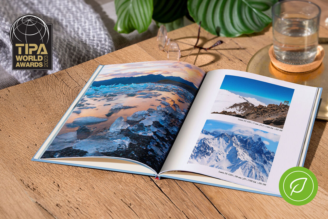 An open CEWE photo book with stunning landscapes on a wooden table. A glass of water and a plant are also visible.