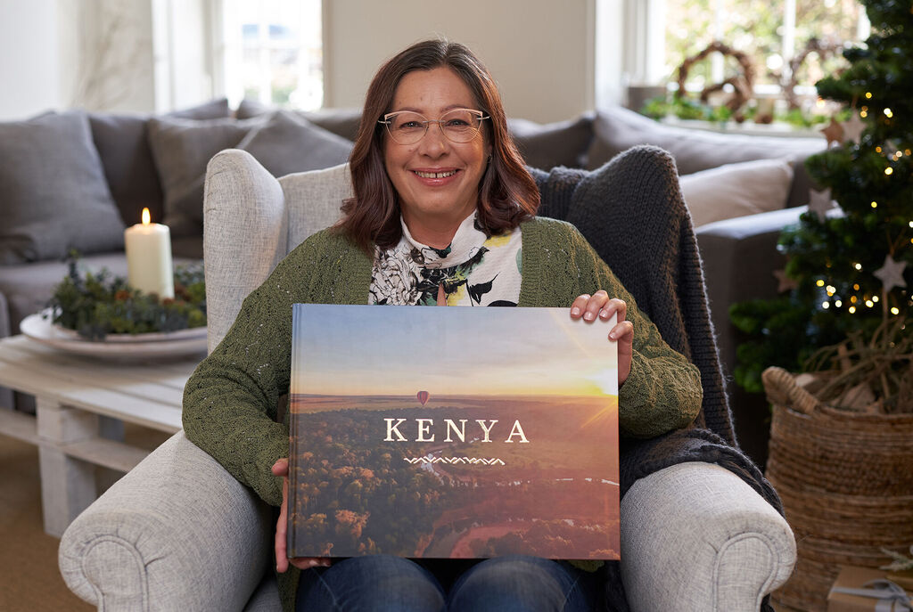 A person holds a CEWE PHOTOBOOK labeled 'KENYA' in hands. In the background is a cozy living room and holiday decor.
