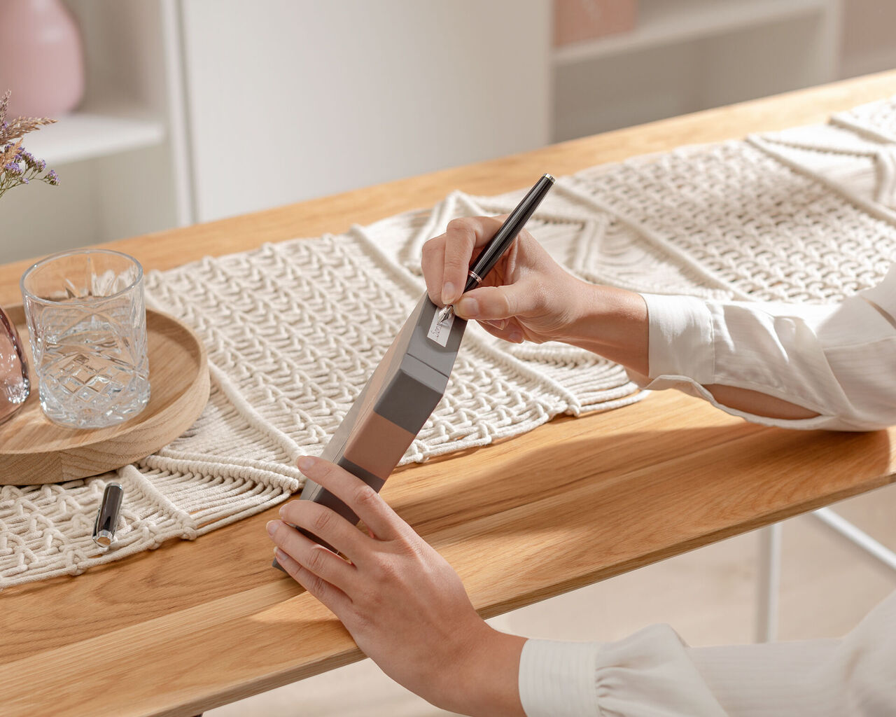 A person holds a gray notebook and writes with a black pen. The table is wooden with decorative elements.