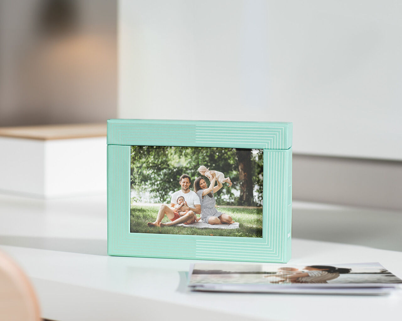 A lovingly designed picture frame with a family photo in a park. In the background, green trees and bright lights are visible. A photo book lies beside it.