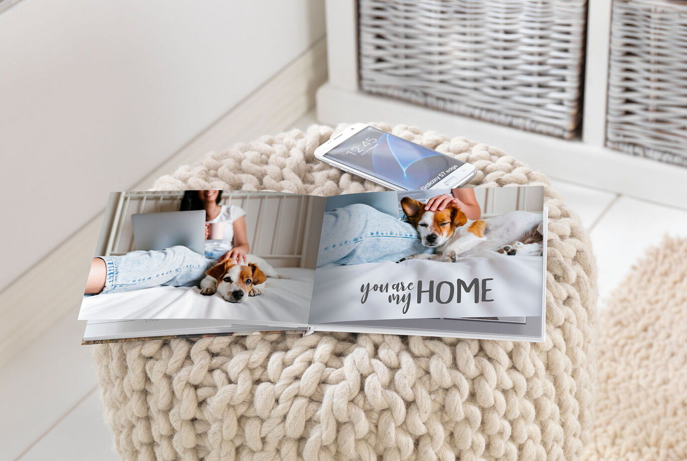 A CEWE PHOTOBOOK with photos and the inscription "you are my HOME" is lying on a stool next to a smartphone. The stool is made of a soft woven material.