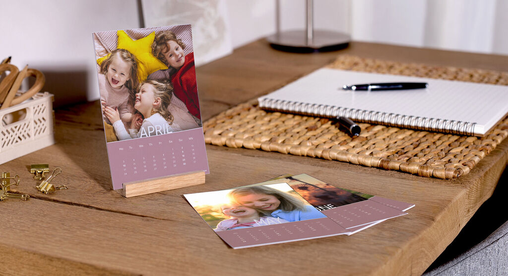 A table with a CEWE photo calendar for April next to photos. The calendar pages show happy moments.
