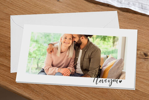 A cheerful couple sits together on a sofa, lovingly hugging and smiling. A lovingly designed envelope with 'Love you' is on the table.