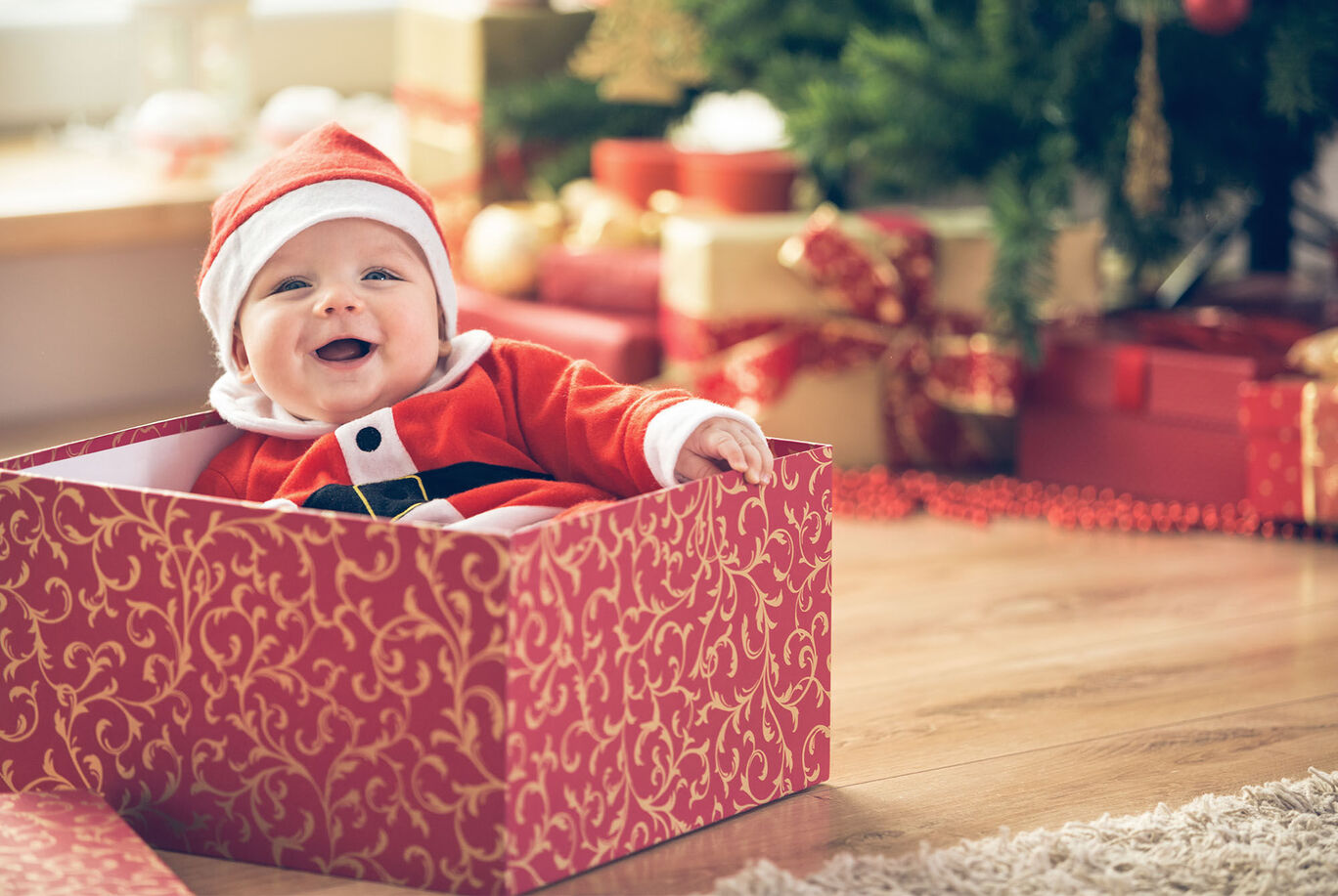 A child in a Santa costume sits in a decorative box in front of a Christmas tree.