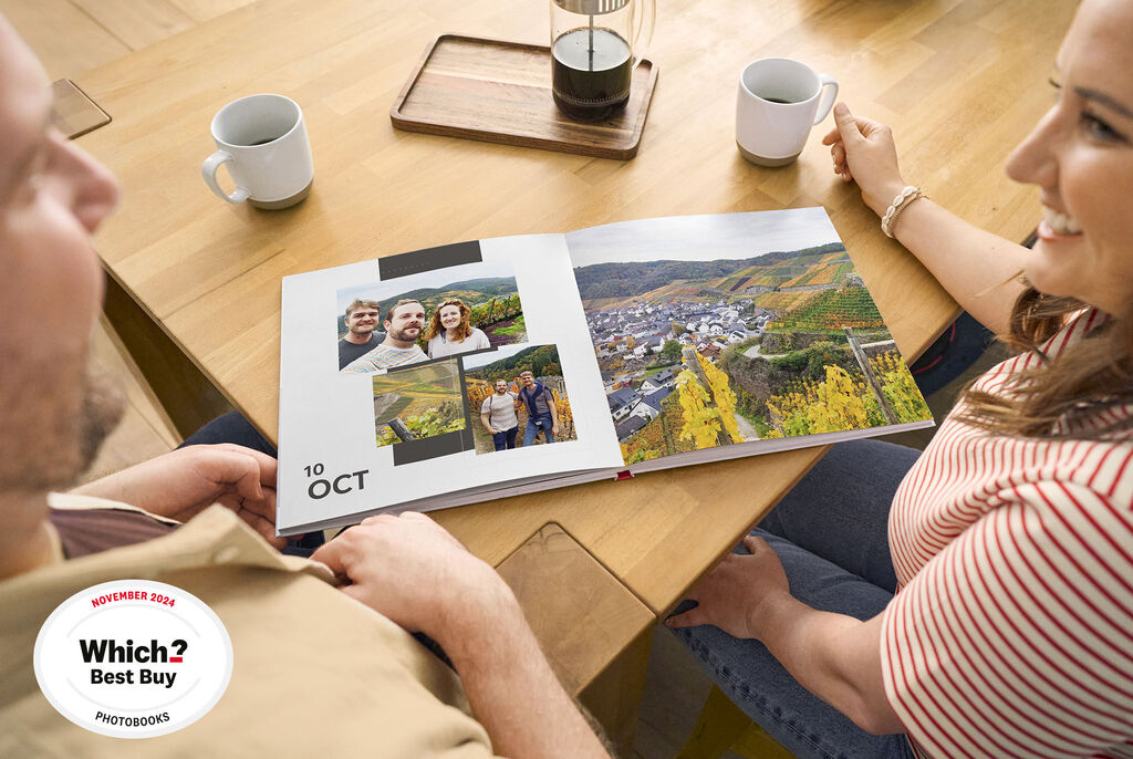 A couple looks at a CEWE photo book featuring their holiday memories at a table with coffee mugs and a lamp.