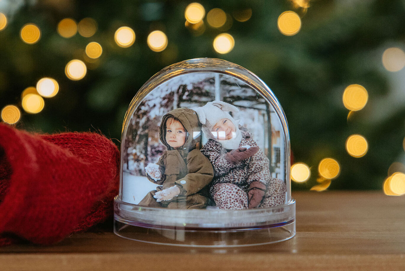 A Christmas ornament with two dolls inside, in front of a decorated tree.