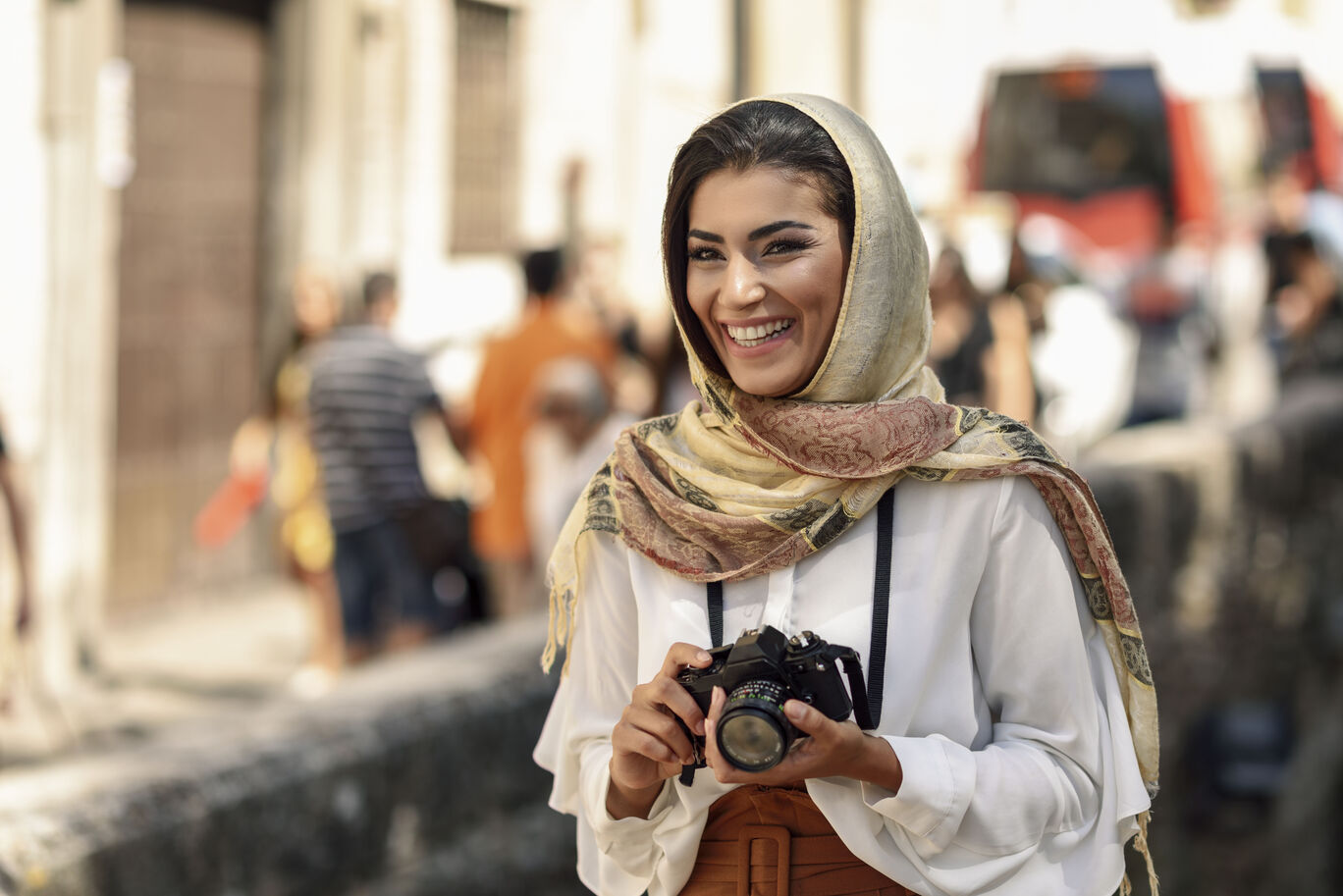 A person holds a camera and wears a scarf, surrounded by people.