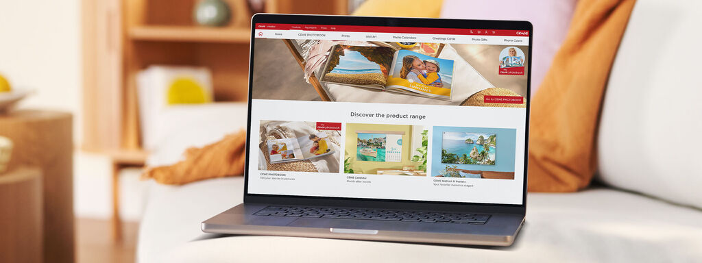 A laptop shows the CEWE website with photo books on a table in a cozy living room.