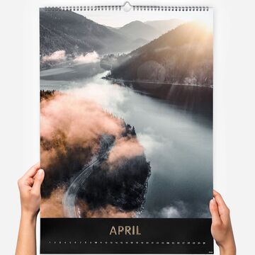 A hand holds a photo calendar with a beautiful landscape for April, showing mountains and a river.