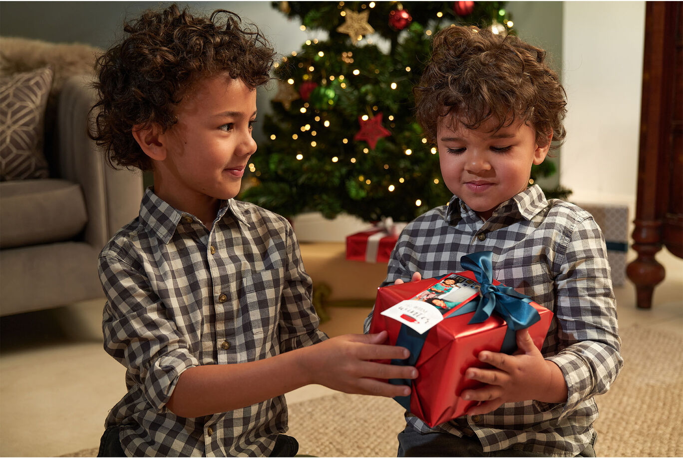 Two children are giving each other a lovingly wrapped gift in front of a festively decorated Christmas tree.