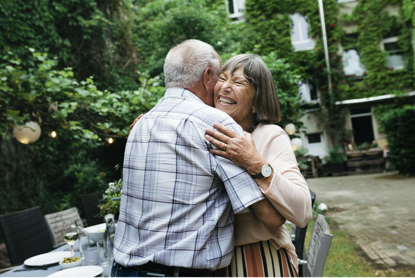 An older couple warmly embraces outdoors, surrounded by green plants and a festive table.